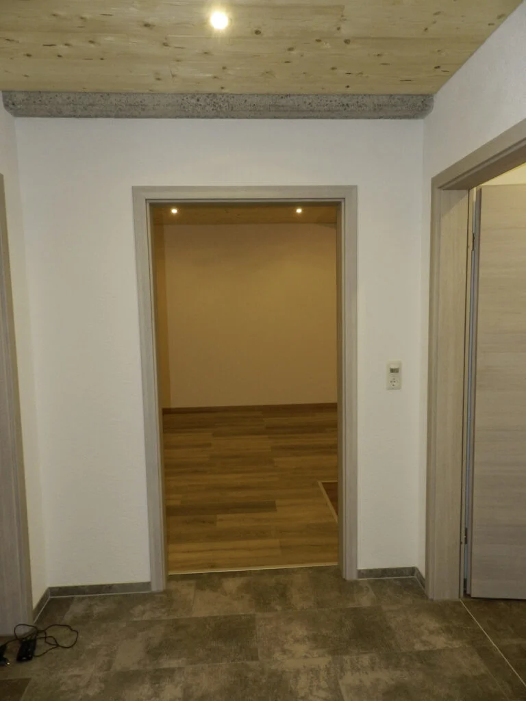 T&uuml;r&ouml;ffnung in einen leeren Raum mit hellem Parkettboden, gedeckten W&auml;nden, Deckenspots und gefliester Schwelle