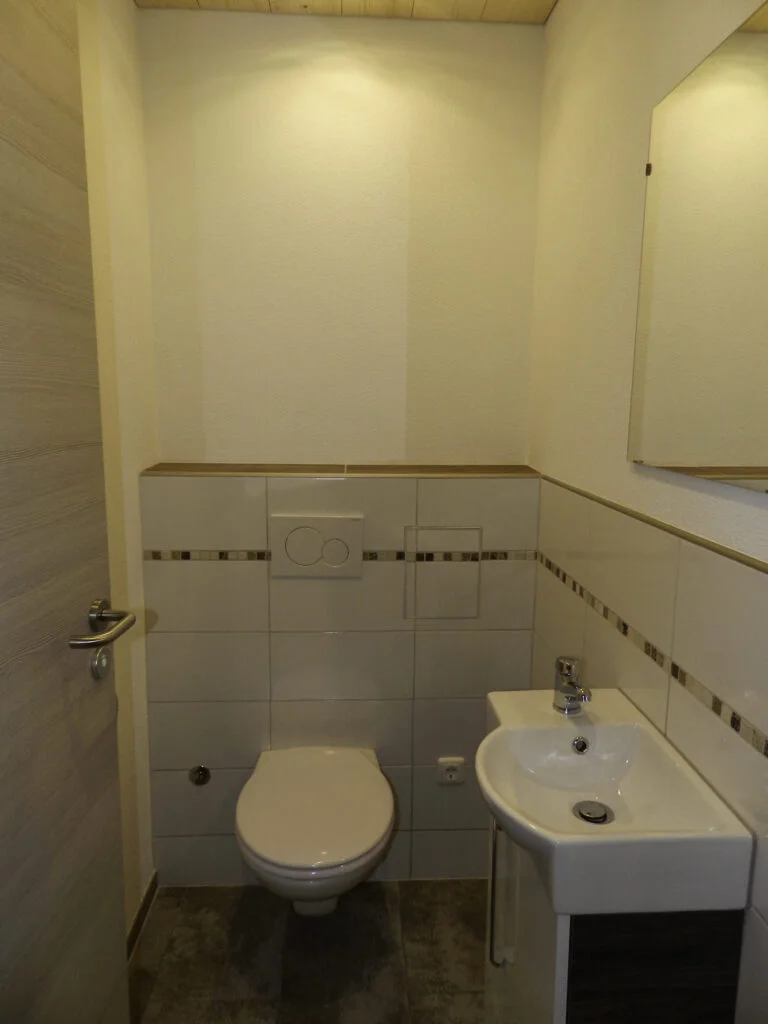 Kleines wei&szlig; gefliestes Badezimmer mit Toilette, kleinem Handwaschbecken, Spiegel und dekorativem Fliesenbord