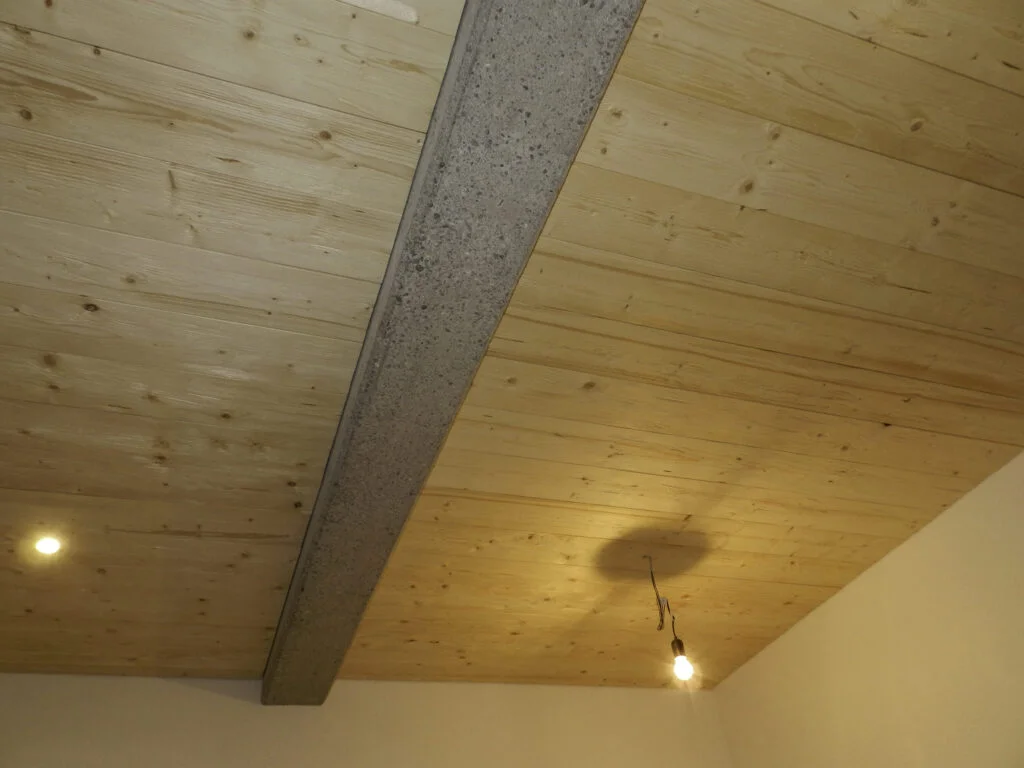 Holzdecke mit hellem Holzfurnier, ein dunkler Deckenbalken und eine nur mit Gl&uuml;hbirne best&uuml;ckte H&auml;ngelampe