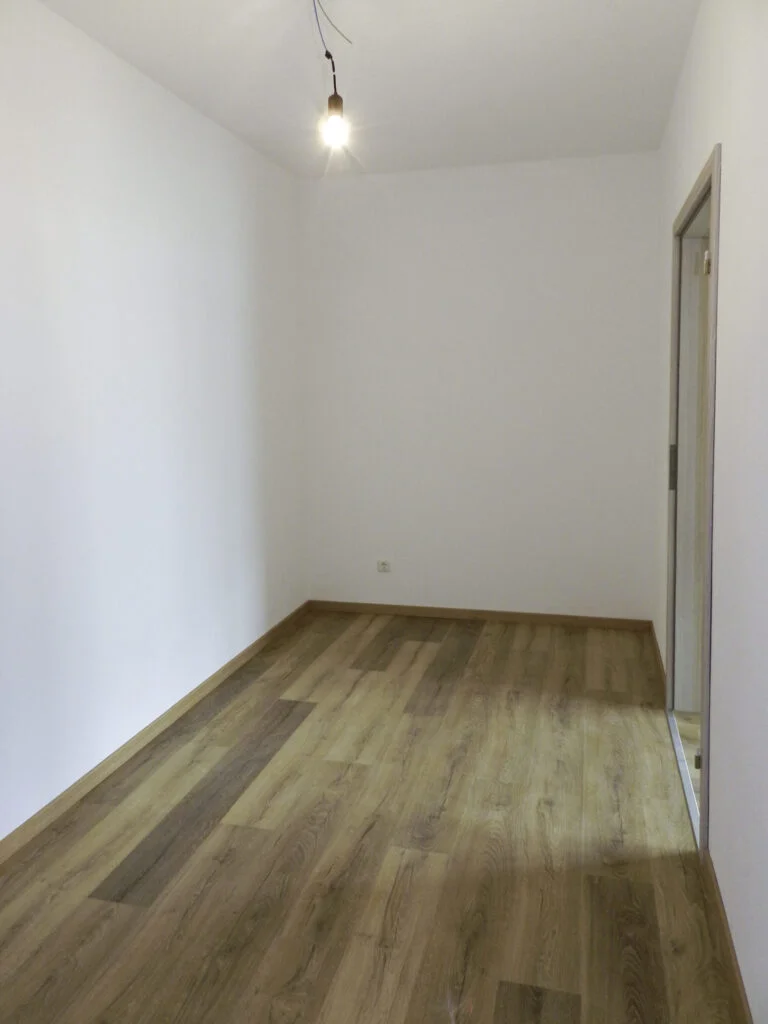 Schmaler leerer Raum mit wei&szlig;en W&auml;nden, Holzboden, Deckenleuchte und T&uuml;r&ouml;ffnung rechts &mdash; in einer Mietwohnanlage in Stanzach.