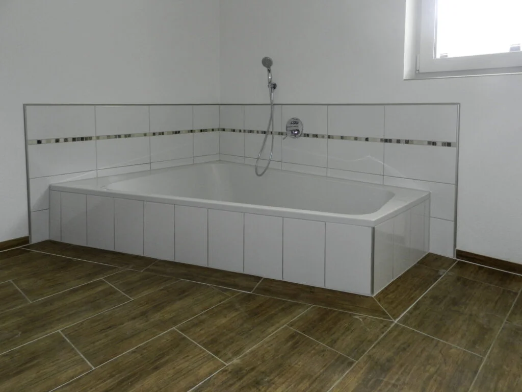 eingebaute wei&szlig;e eckige Badewanne mit gefliertem Rand, Bord&uuml;re, Boden in Holzoptik und kleinem Fenster; Mietwohnung