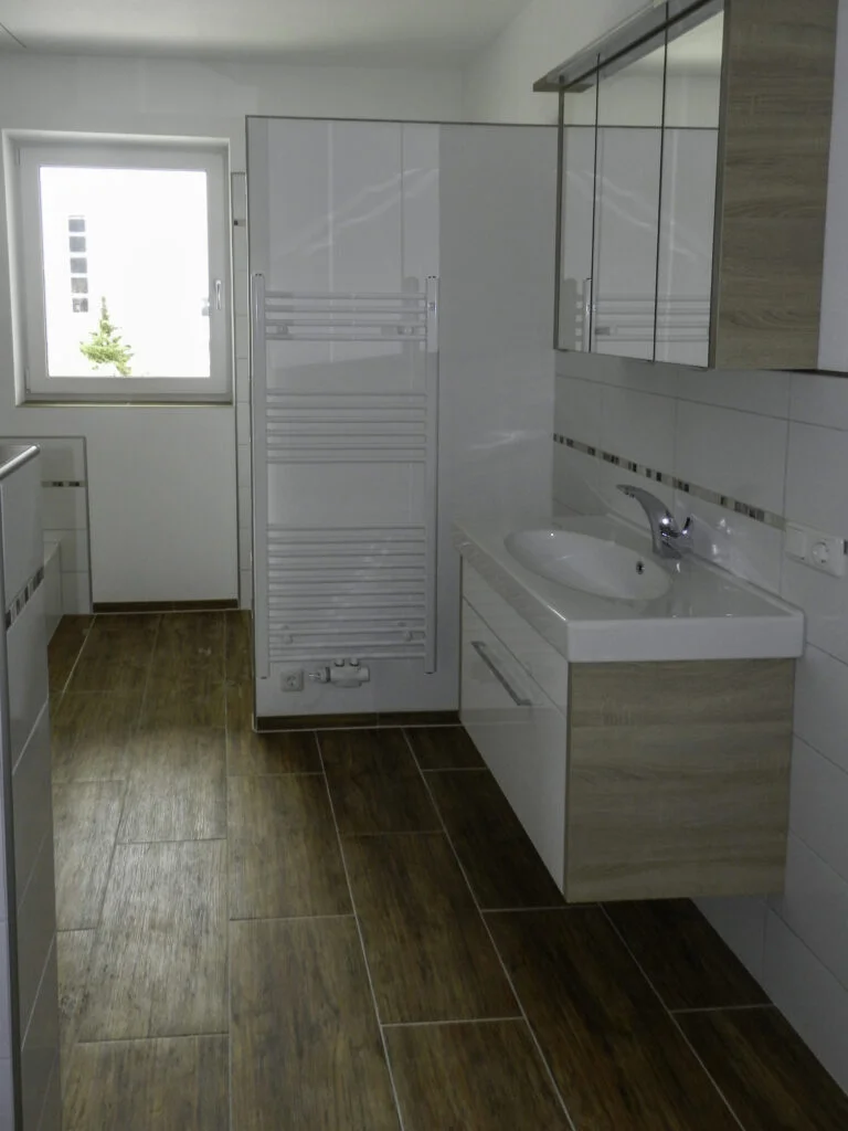 Kompaktes Badezimmer mit h&auml;ngendem Waschtisch und Spiegelschrank, vertikalem Handtuchheizk&ouml;rper, Fenster und Holzoptikboden.