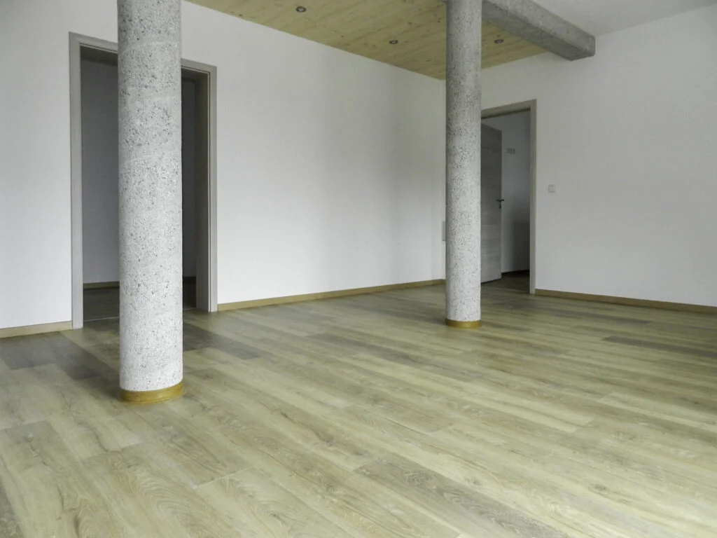Leerer Wohnraum in Mietwohnanlage Stanzach mit zwei Betons&auml;ulen, wei&szlig;en W&auml;nden und hellem Laminatboden