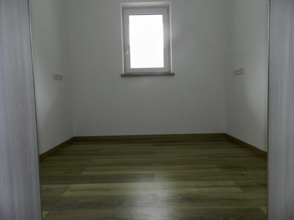 Kleines leeres Zimmer in Mietwohnung, wei&szlig;e W&auml;nde, schmales Fenster mittig, dunkler Laminatboden, keine M&ouml;bel.