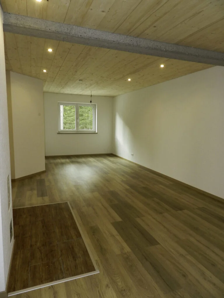 Leerer Wohnraum mit wei&szlig;en W&auml;nden, dunkelbraunem Laminatboden, sichtbaren Deckenbalken mit Einbauleuchten und kleinem Fenster