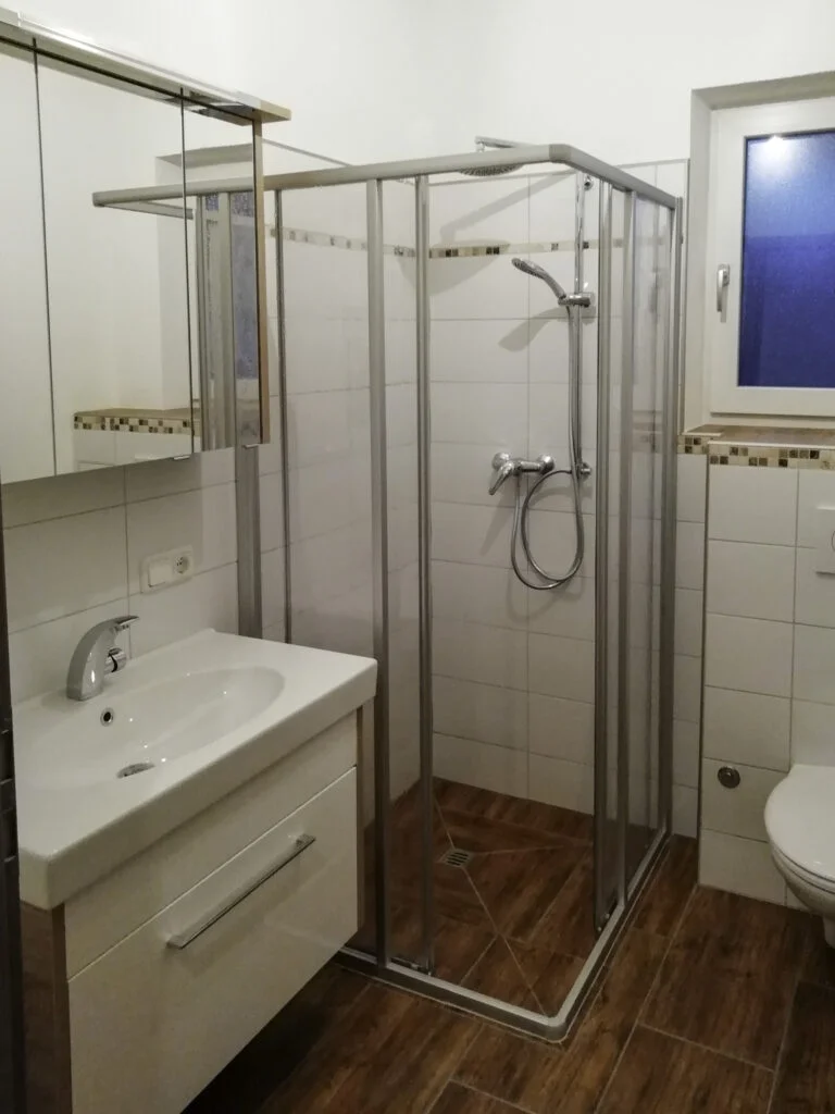 Kleines wei&szlig;es Badezimmer mit Dusche, Glasduscht&uuml;r, wei&szlig;em Waschtisch mit Schubladen, WC und Holzoptik-Boden.