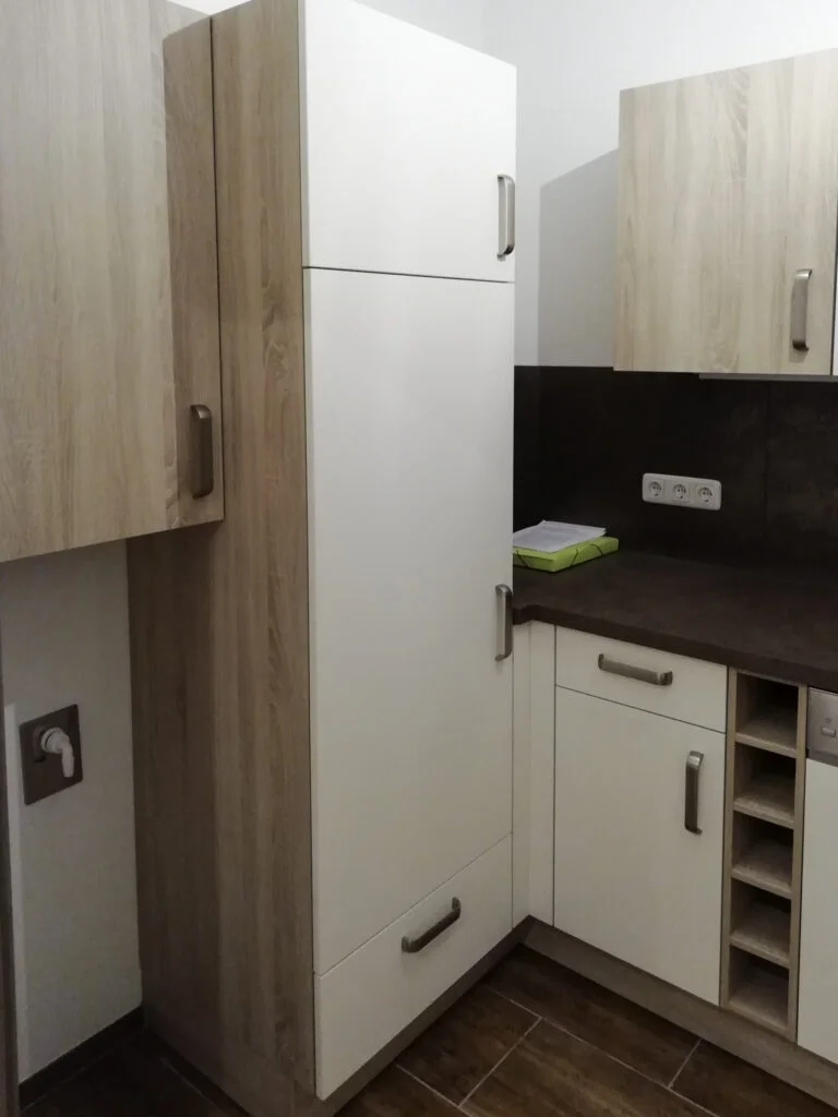 Kleine Einbauk&uuml;che in Mietwohnung mit hohem wei&szlig;en K&uuml;hlschrank, hellen Holzschr&auml;nken und dunkler Arbeitsplatte.