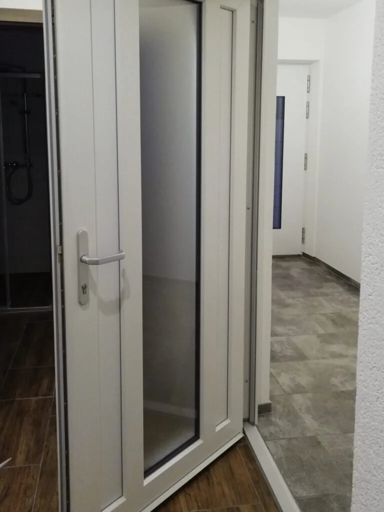 Ge&ouml;ffnete wei&szlig;e Glast&uuml;r mit Griff in schmalem Flur einer Mietwohnung, heller Bodenbelag und wei&szlig;e W&auml;nde.