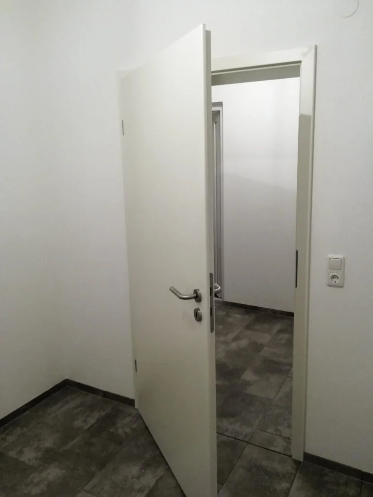Enger Wohnungsflur mit ge&ouml;ffneten wei&szlig;en T&uuml;ren, helle W&auml;nde, graue Bodenfliesen, T&uuml;rgriff und Lichtschalter
