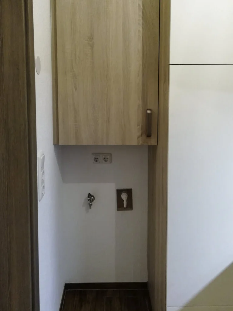 Nische in Wohnung mit Wasseranschluss, Abfluss und Steckdose unter einem h&ouml;lzernen Wandschrank, wei&szlig;e Wand