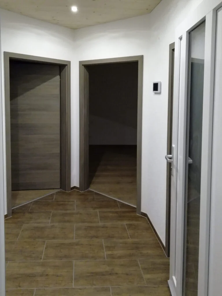 Flur mit braunem Boden, wei&szlig;en W&auml;nden, zwei offenen T&uuml;ren links und einer T&uuml;r mit Glaseinsatz rechts