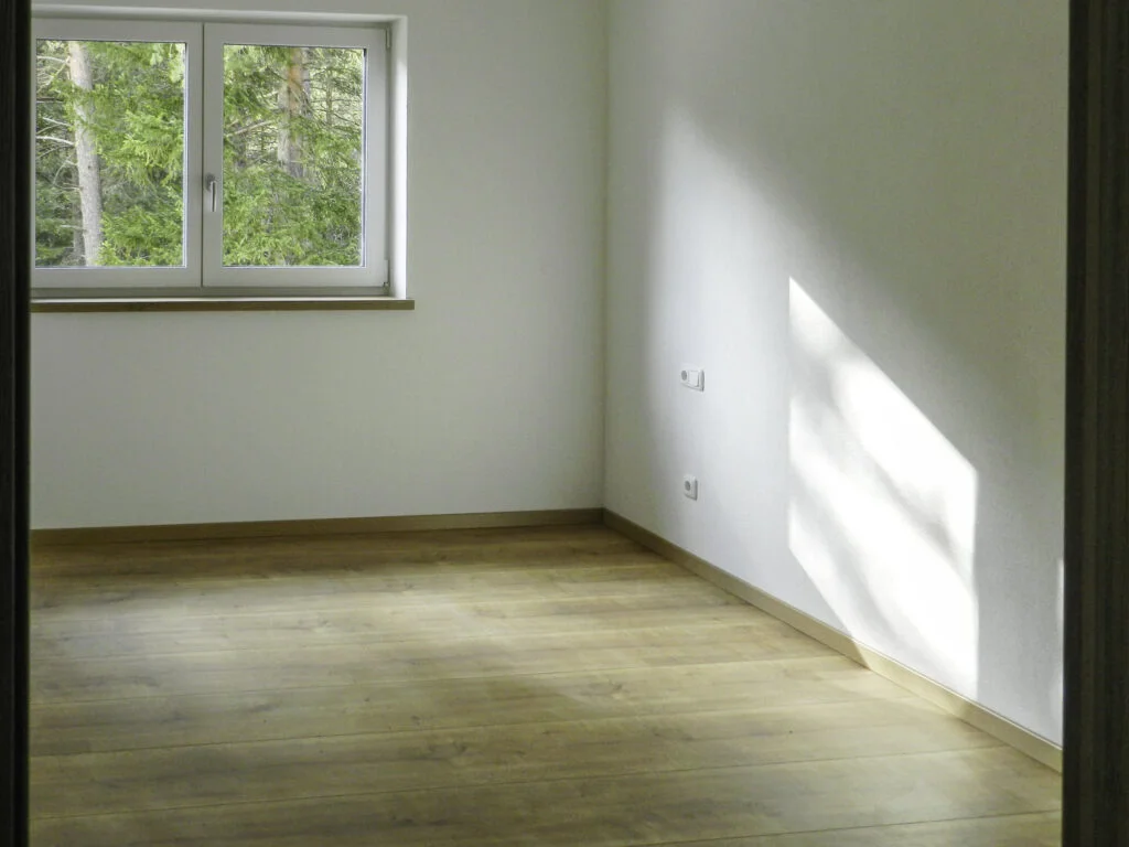 Leeres Zimmer einer Mietwohnung mit Fenster, hellem Holzboden, wei&szlig;en W&auml;nden und Lichtstreifen an der Wand.