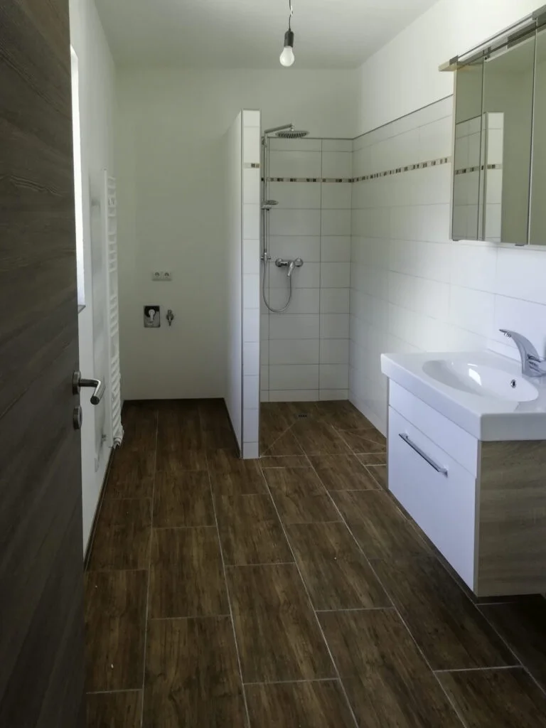 Badezimmer mit wei&szlig;em Waschtisch und Spiegelschrank, offener Duschbereich und holzfarbenen Bodenfliesen.