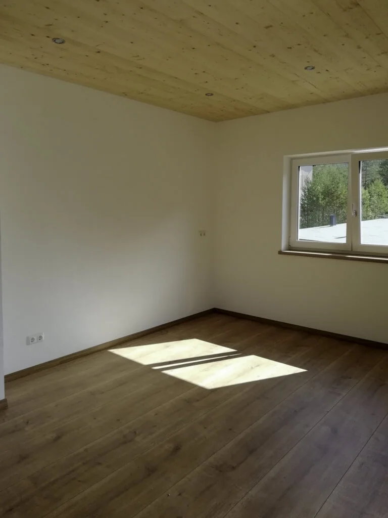 Leerer Wohnraum mit hellem Holzboden, Holzdecke, wei&szlig;en W&auml;nden und Fenster mit Heizk&ouml;rper; Sonnenlicht f&auml;llt auf den Boden.