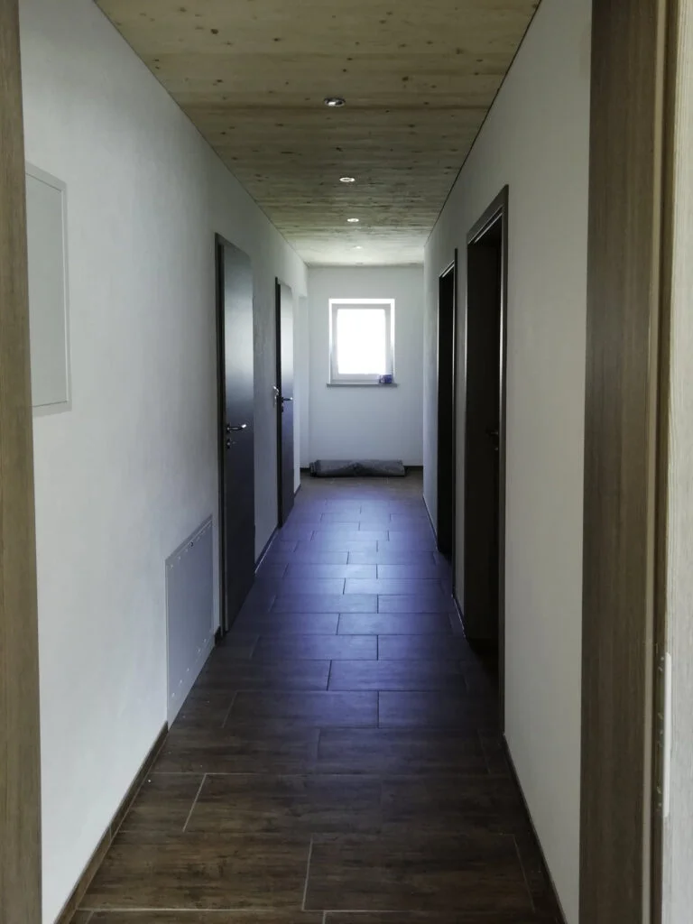 Schmaler Flur einer Mietwohnanlage mit dunklem Laminatboden, wei&szlig;en W&auml;nden, mehreren T&uuml;ren und Fenster am Ende