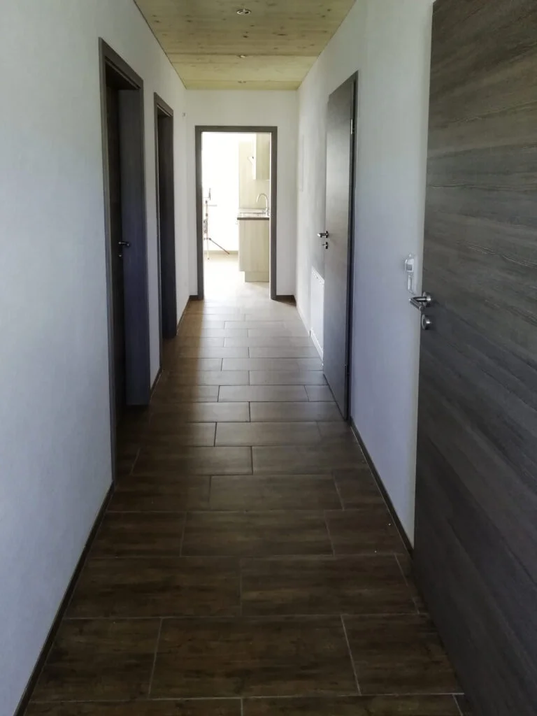 Flur einer Wohnung mit dunklem Laminatboden, wei&szlig;en W&auml;nden, mehreren T&uuml;ren links und rechts und heller &Ouml;ffnung am Ende.