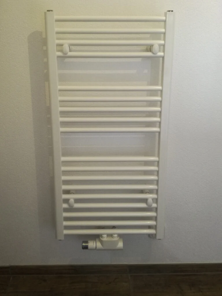 Wei&szlig;er wandmontierter Handtuchheizk&ouml;rper mit waagerechten Streben an heller Wand, Thermostatventil unten