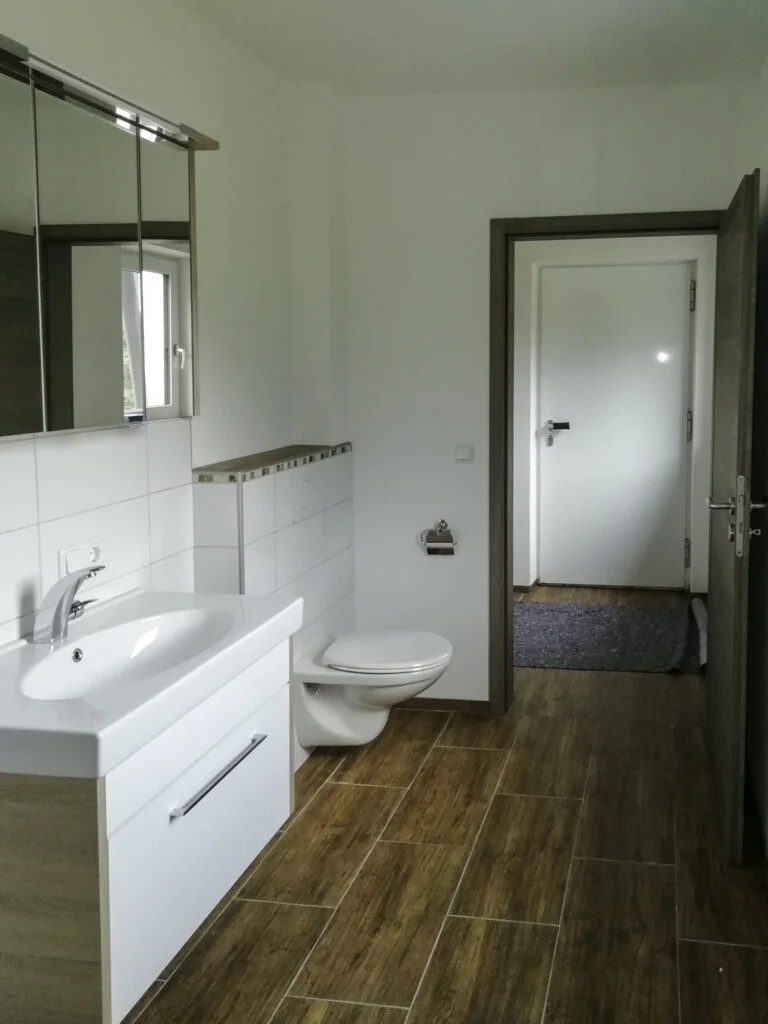 Badezimmer einer Mietwohnung mit Waschbecken und Unterschrank, Spiegel, Toilette, Wandregal und Holzboden; offene T&uuml;r zum Flu