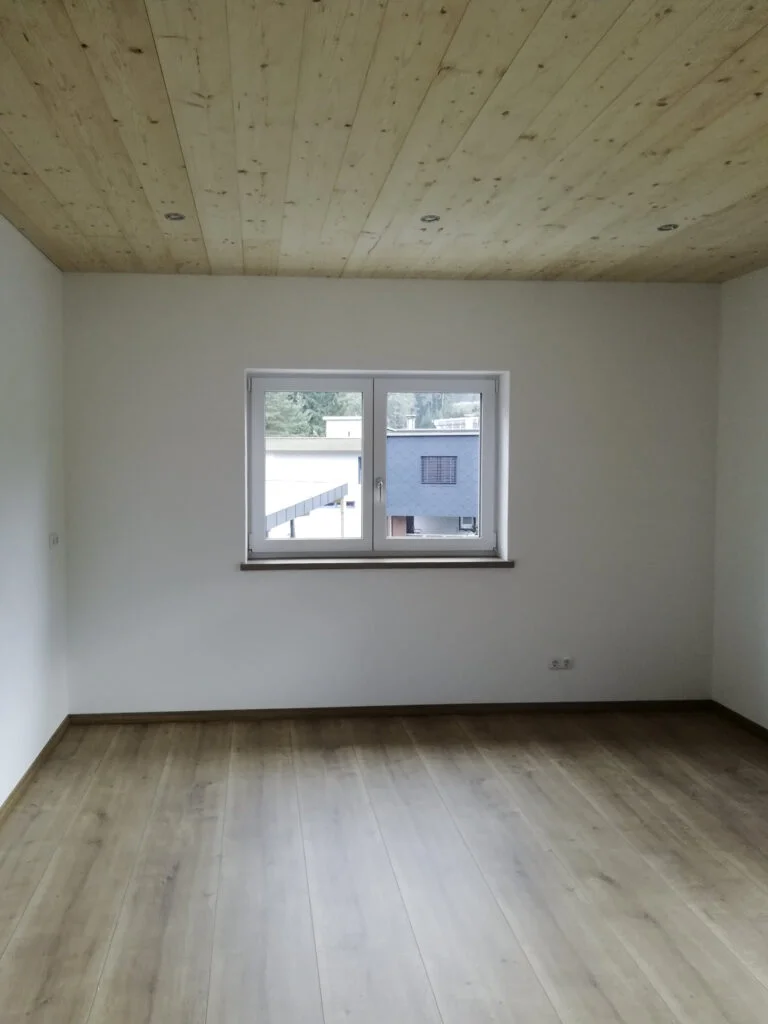 Leerer Wohnraum mit hellem Holzfu&szlig;boden, wei&szlig;en W&auml;nden und Holzdecke; mittiges Fenster mit Blick auf Nachbargeb&auml;ude; Heizk&ouml;rp