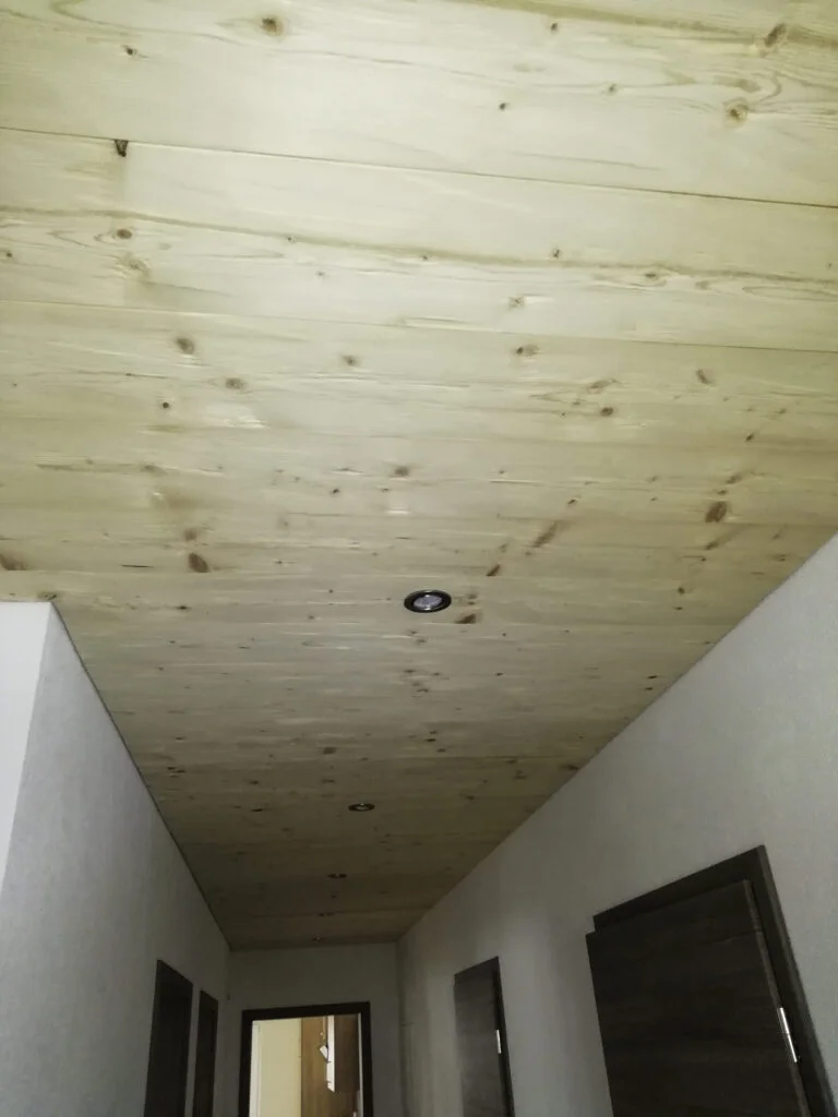 Flur mit wei&szlig;en W&auml;nden, hellen T&uuml;rrahmen und einer sichtbaren Holzdecke mit Knoten und offener Lampenfassung