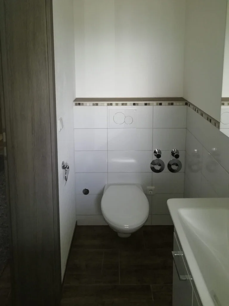 Badezimmer mit wandh&auml;ngendem WC, Waschbecken rechts, wei&szlig;e Fliesen mit Bord, sichtbare Rohranschl&uuml;sse, dunkler Boden