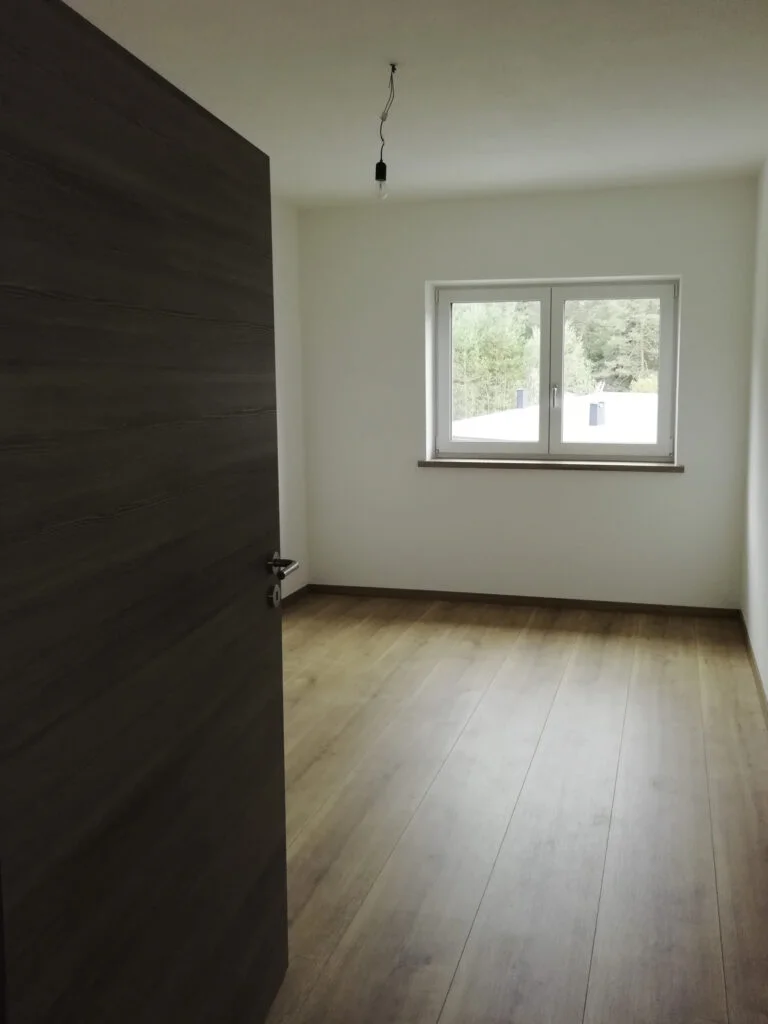 Leerer Raum in Mietwohnung, wei&szlig;e W&auml;nde, Laminatboden, halb offene dunkle T&uuml;r, ein Fenster mit Gr&uuml;nblick, nackte Lampe.