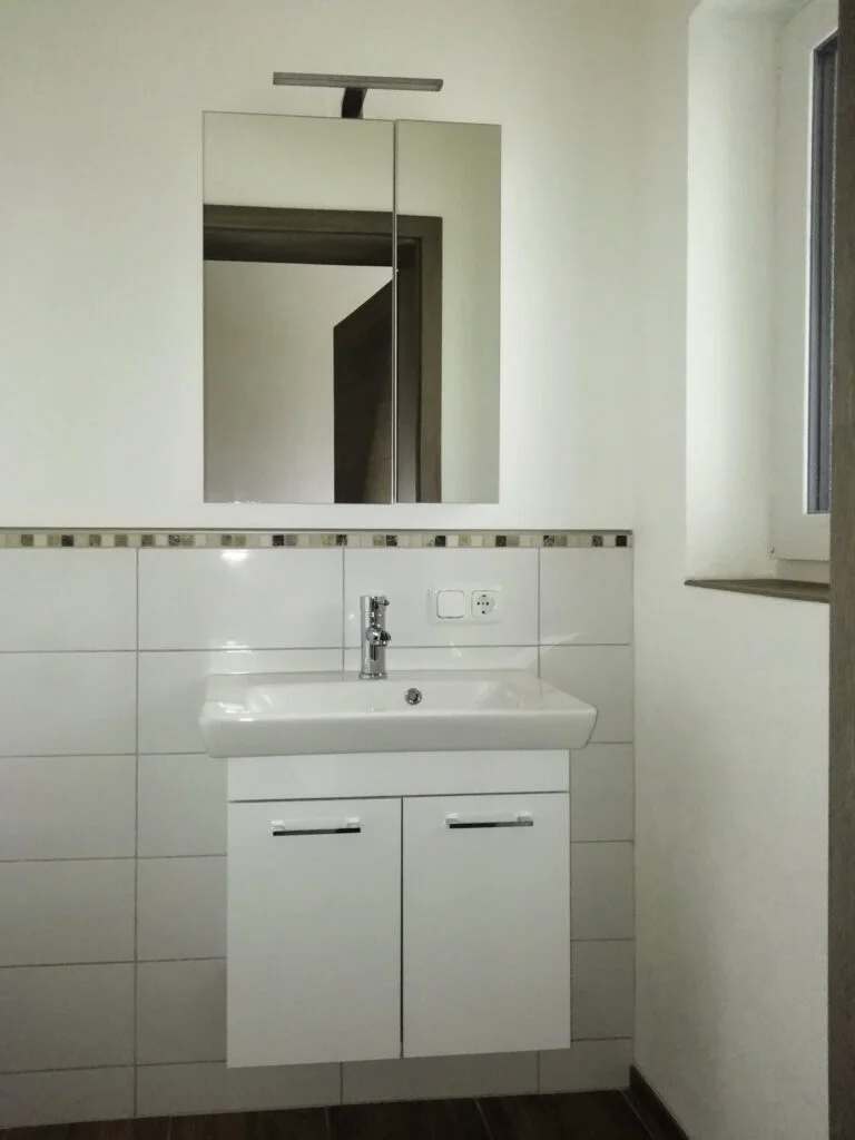 kleines Badezimmer der Mietwohnung mit wei&szlig;em Waschbecken und Unterschrank, Wandspiegel, wei&szlig;e Fliesen und kleines Fenster