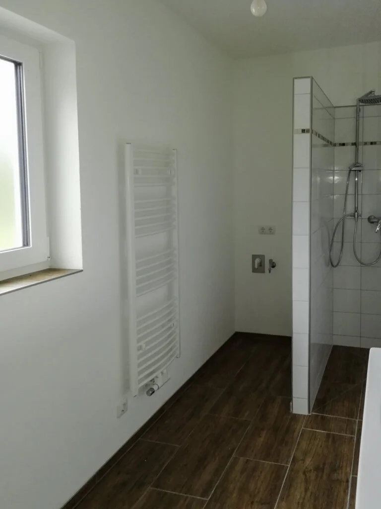 Badezimmer in Mietwohnung: Fenster, Wandheizk&ouml;rper, Duschbereich mit Glasabtrennung und braunem Holzboden.