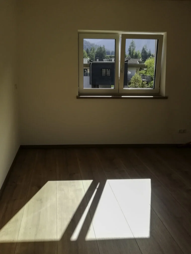 Leerer Raum mit hellem Holzfu&szlig;boden, Sonneneinfall auf dem Boden; Fenster mit Blick auf Nachbarh&auml;user und B&auml;ume.